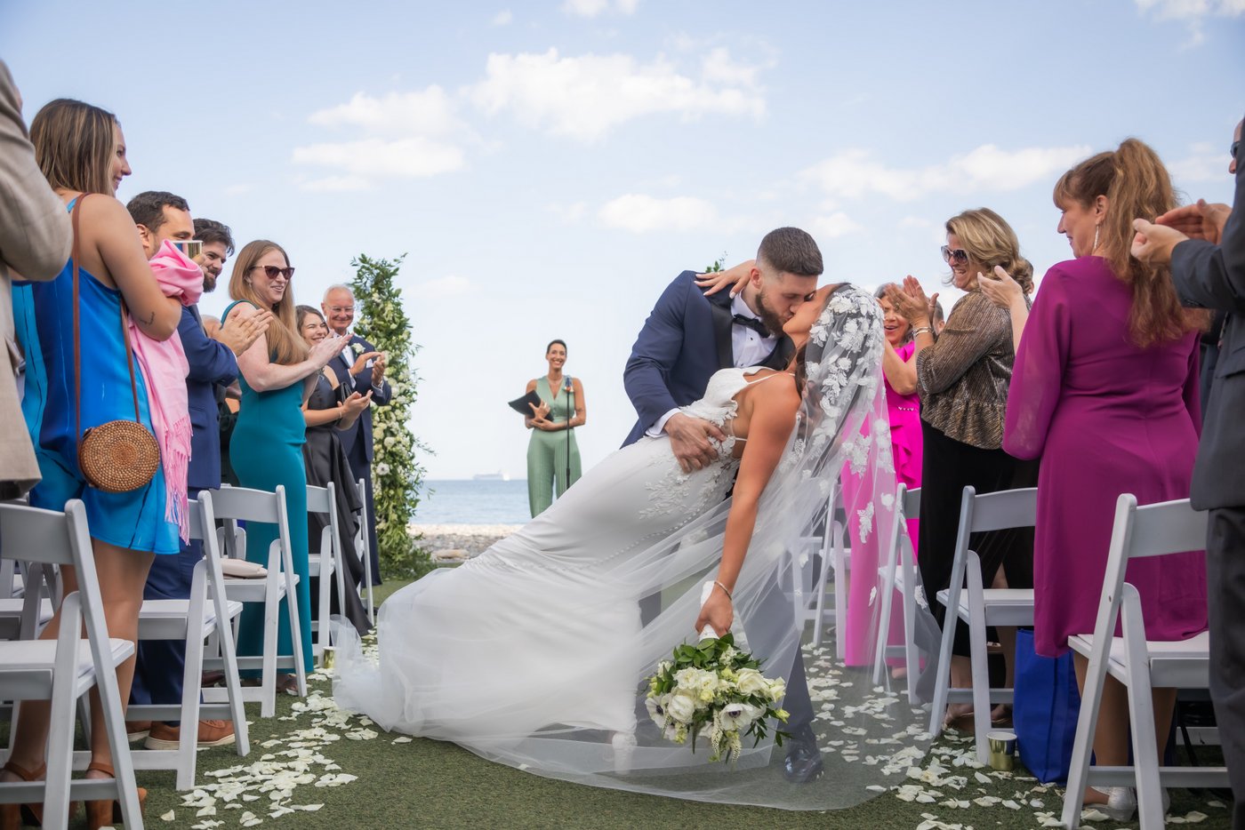 The groom dips and kisses the bride as wedding guests stand and clap during their wedding ceremony recessional at their Oceanview of Nahant wedding