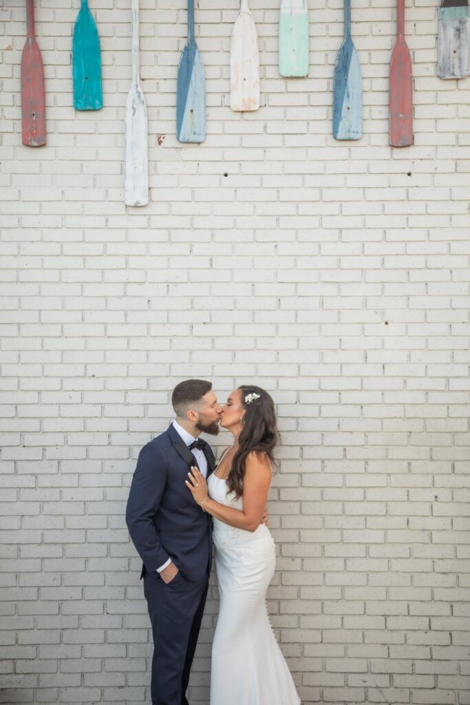 The bride and groom kiss by the gray wall with colorful hanging ores above them during their Oceanview of Nahant wedding portraits