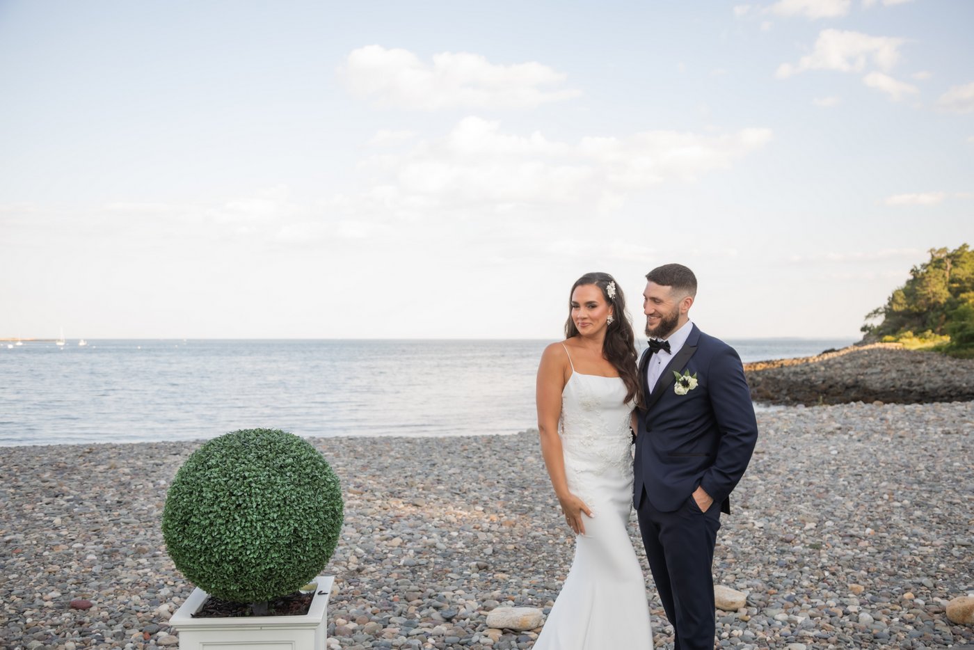 The groom looks lovingly at the bride while the bride looks at the camera and smiles with the ocean behind them and the topiary beside them at an Oceanview of Nahant wedding.