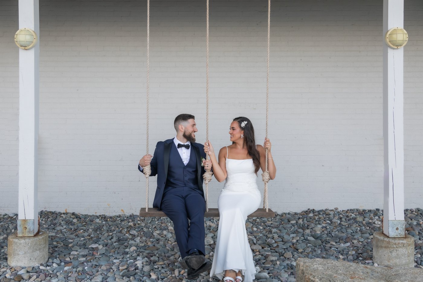 The bride and groom sit on the swings and smile at each other during their wedding portraits at Oceanview of Nahant