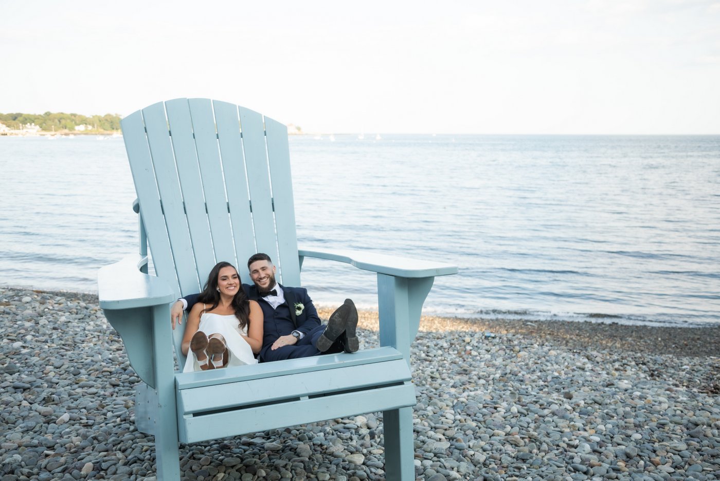 The bride and groom relax on the giant blue Adirondack chair by the water during wedding portraits at Oceanview of Nahant wedding venue in Massachusetts.