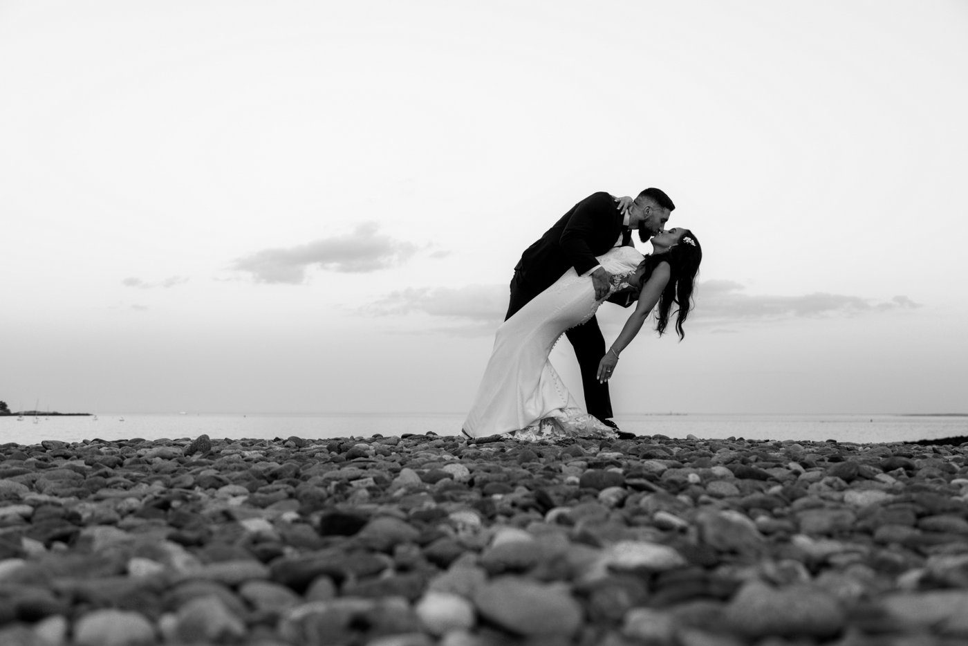 A black and white photo of the groom dipping and kissing the bride at sunset during portraits at Oceanview of Nahant, a popular coastal wedding venue on the North Shore of MA.