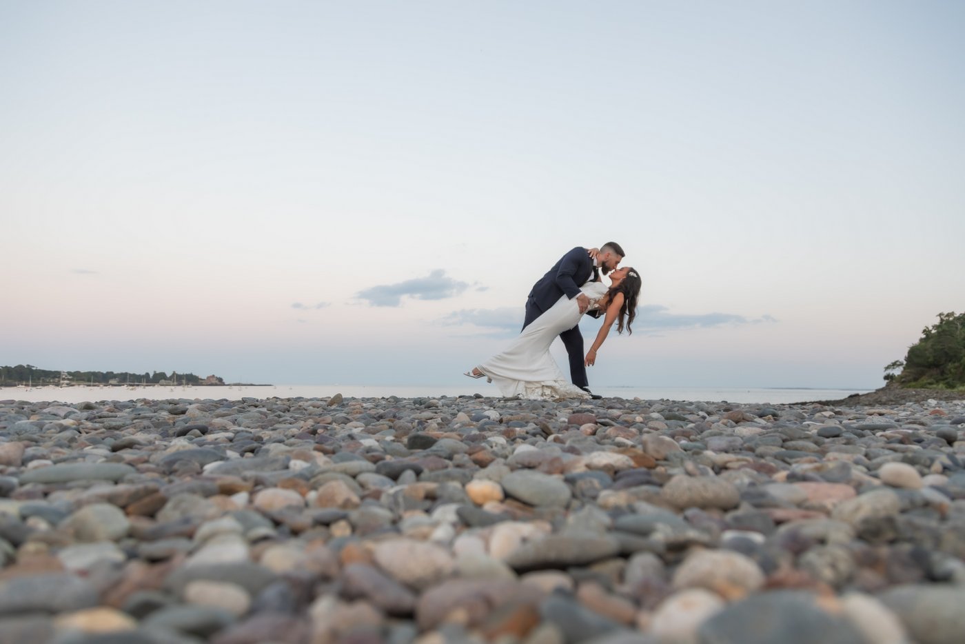 The groom dips and kisses the bride on the pebbled shoreline of the beach at sunset at the Oceanview of Nahant wedding, photographed by creative Boston wedding photographers Spagnolo Photography