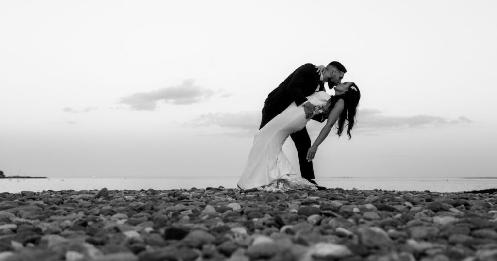 A black and white photo of the groom dipping and kissing the bride at sunset during portraits at Oceanview of Nahant, a popular coastal wedding venue on the North Shore of MA.
