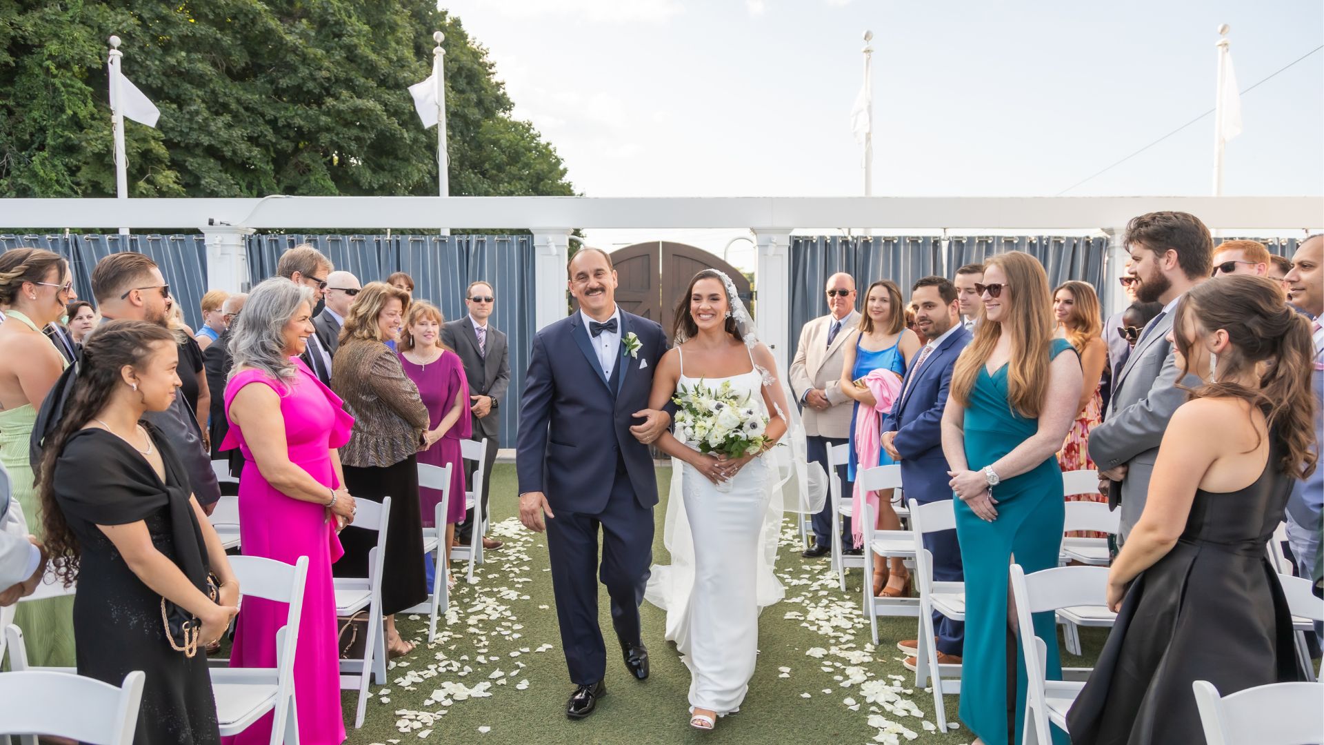The bride and her father smile as they walk down the aisle overlooking the water at an Oceanview of Nahant wedding in coastal Massachusetts.