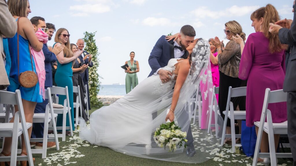 An Oceanview of Nahant wedding romantic moment, when the groom dips and kisses the bride in the aisle during their wedding ceremony exit in front of their wedding guests and the ocean behind them.