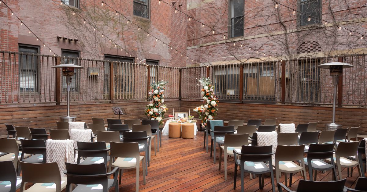 The patio ceremony space decorated with a floral altar, a table with sweets, a pair of leather stools for the bride and groom, and cozy throws on chairs for the guests, ahead of an elegant SRV Boston wedding.