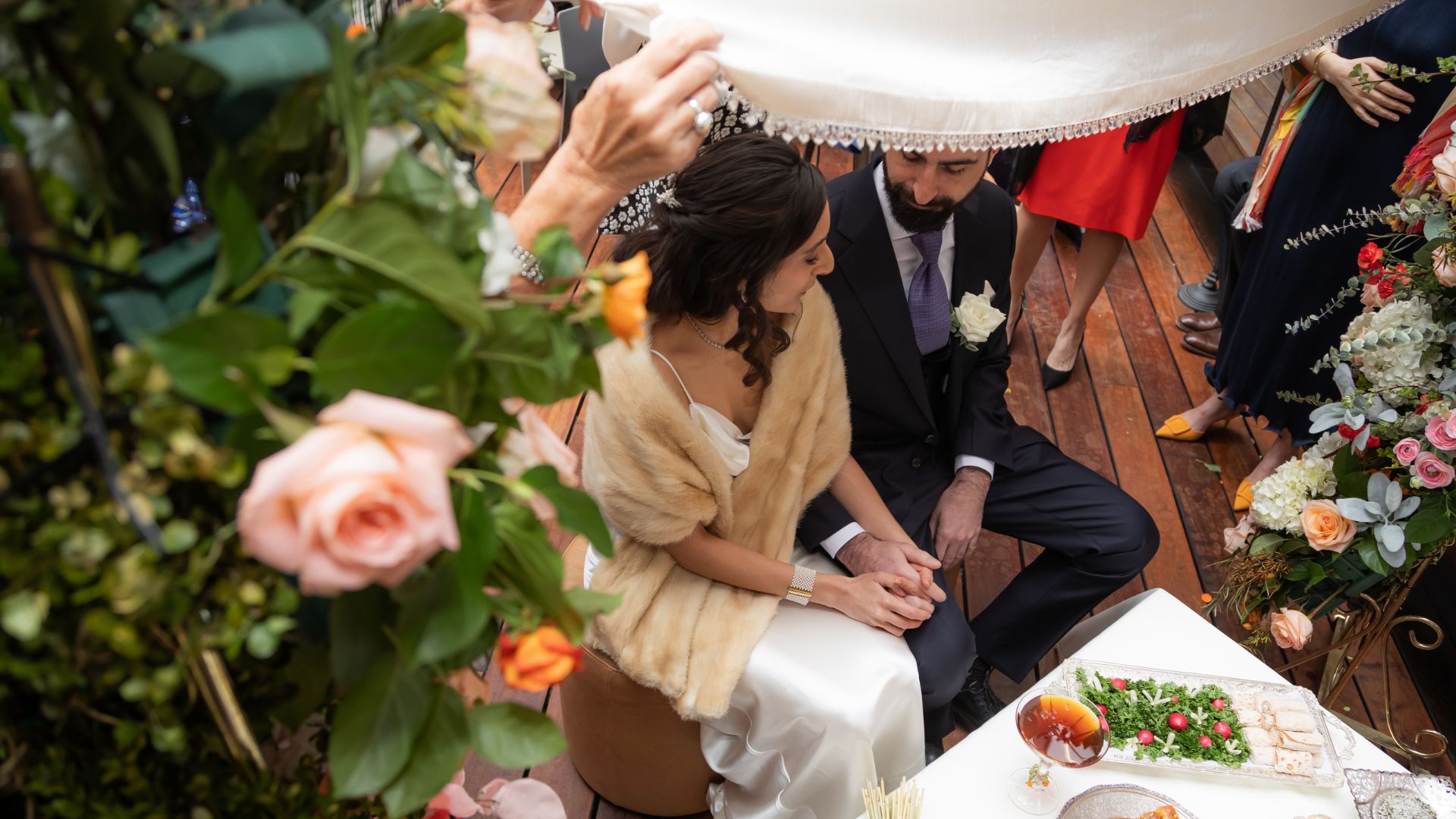 Bride and groom sit under a white canopy in front of the floral altar and their Persian sofreh aghd table during their SRV Boston wedding ceremony.