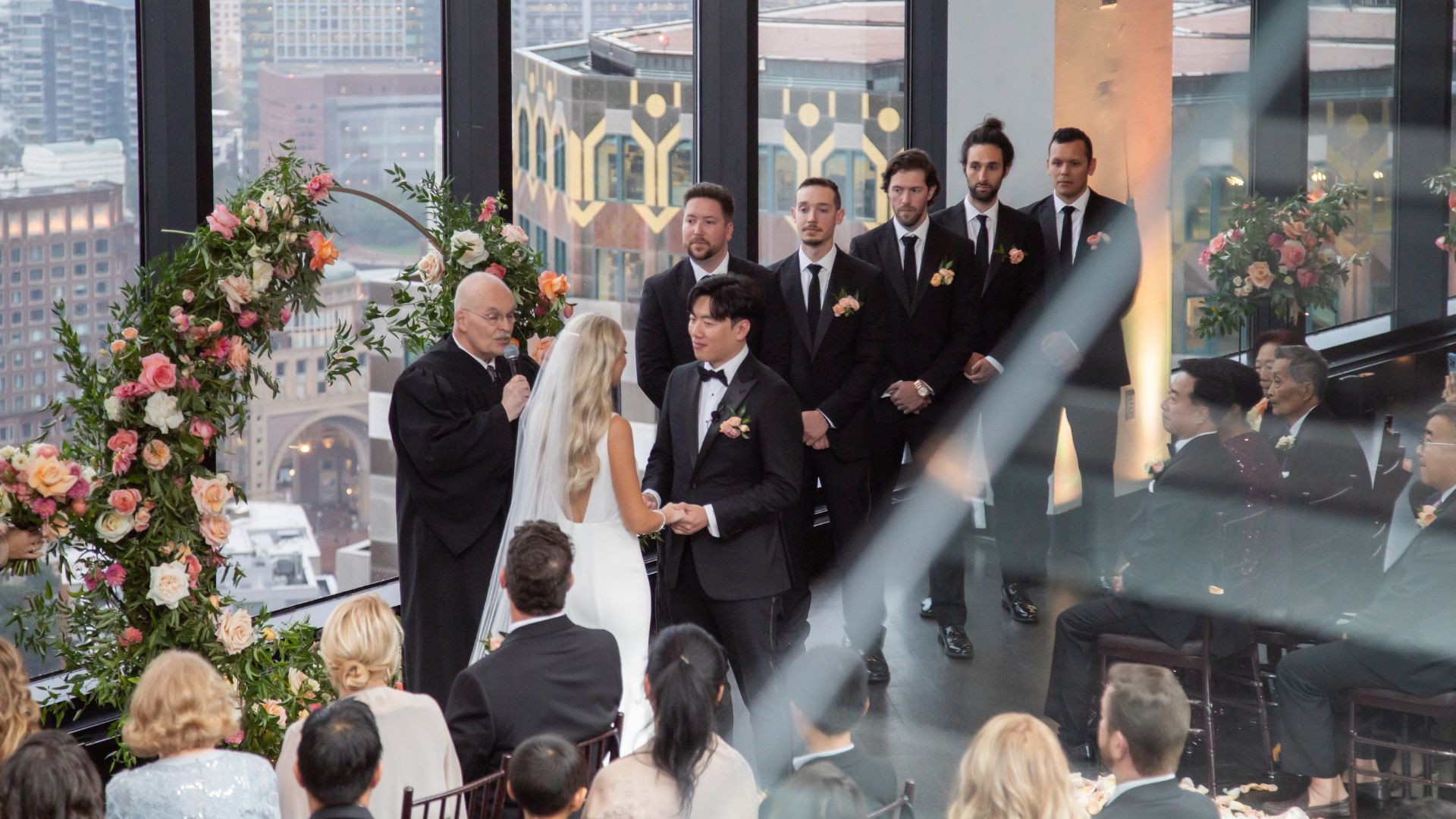 The bride and groom hold hands during their State Room Boston wedding ceremony with Boston's skyline behind them in the Great Room of State Room Boston: A Longwood Venue.