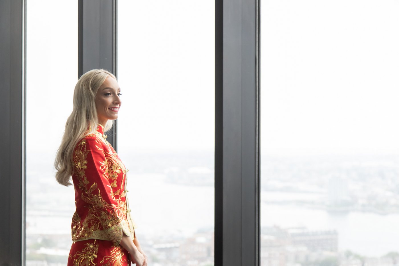 The bride in her red and gold Qun Kwa standing in front of the tall glass windows a the her wedding venue State Room in Boston, with the city skyline behind her