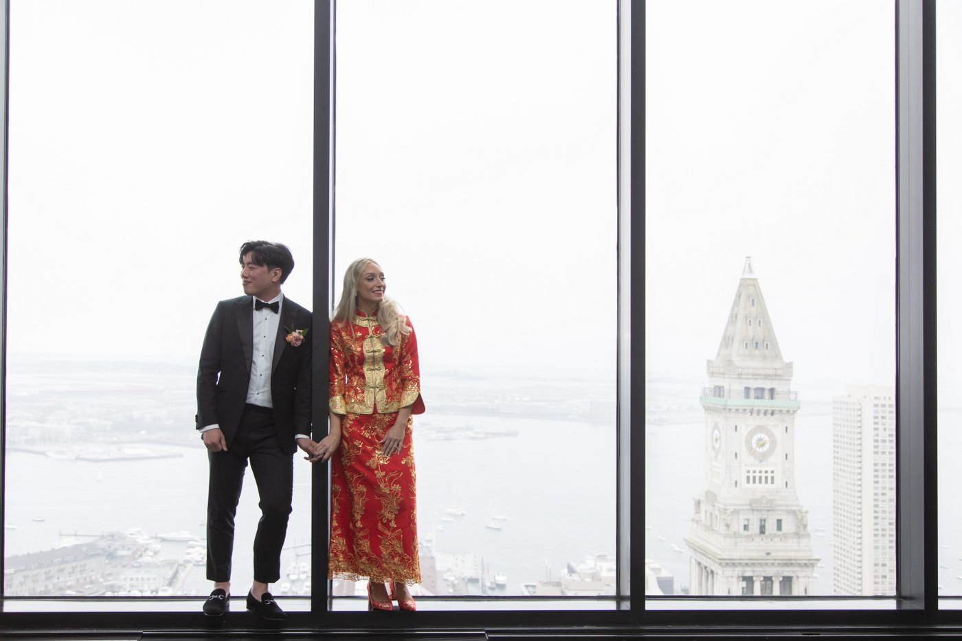 The bride in her red and gold Qun Kwa and the groom in his tux stand in front of the iconic glass windows of the State Room in Boston, with the city skyline behind them on their wedding day