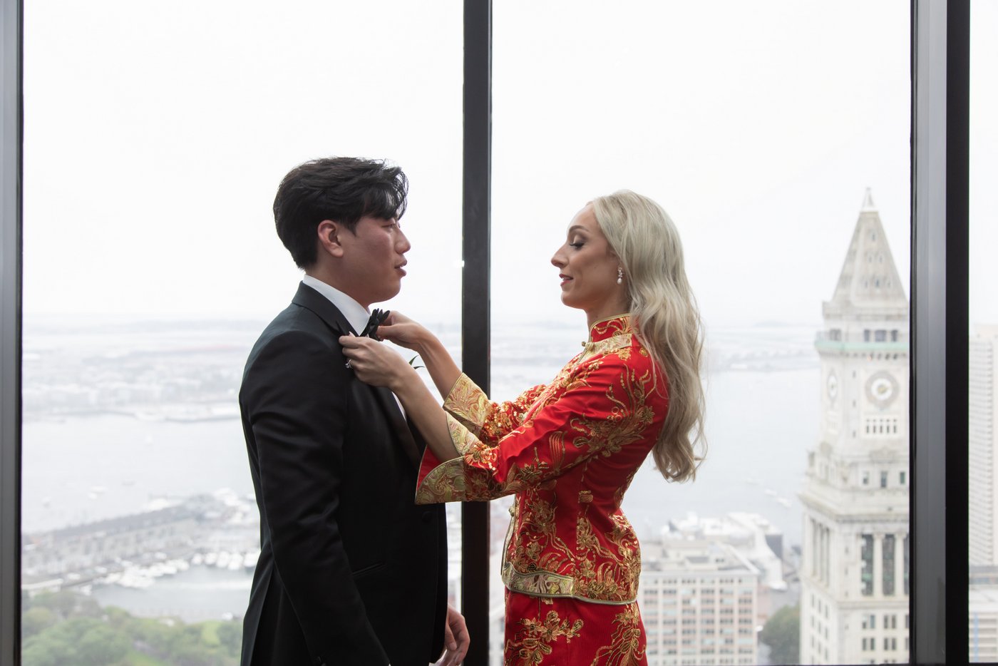 The bride in her red and gold Qun Kwa adjusts the groom's bowtie in front of the iconic glass windows of the State Room in Boston on their wedding day, with the city skyline behind them