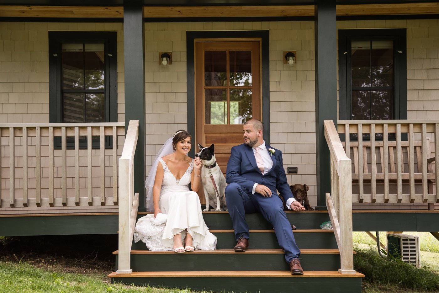 A bride and a groom and their two dogs sit on the steps of a little cabin during their rustic barn wedding in Massachusetts