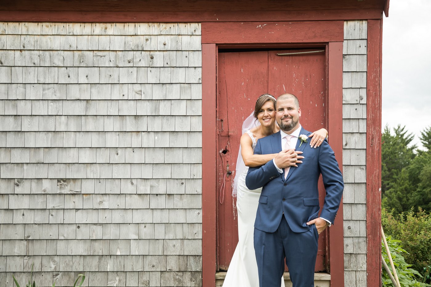 A bride and groom smile at the camera while hugging in front of a rustic gray cabin with red trim