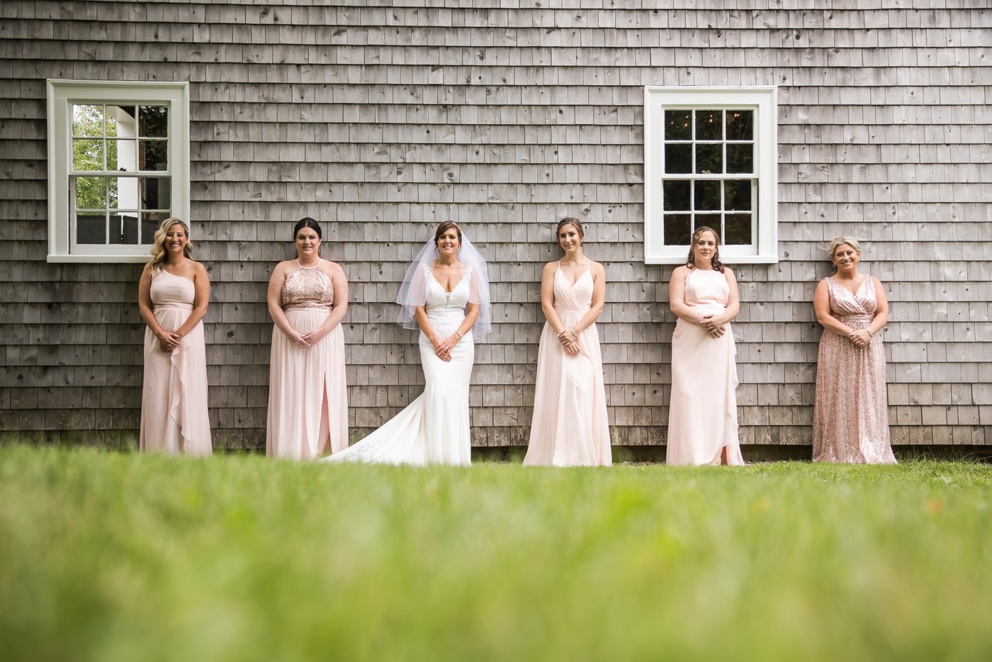 The bride and her bridesmaids wearing pink dresses stand against a rustic gray cabin during a Massachusetts barn wedding