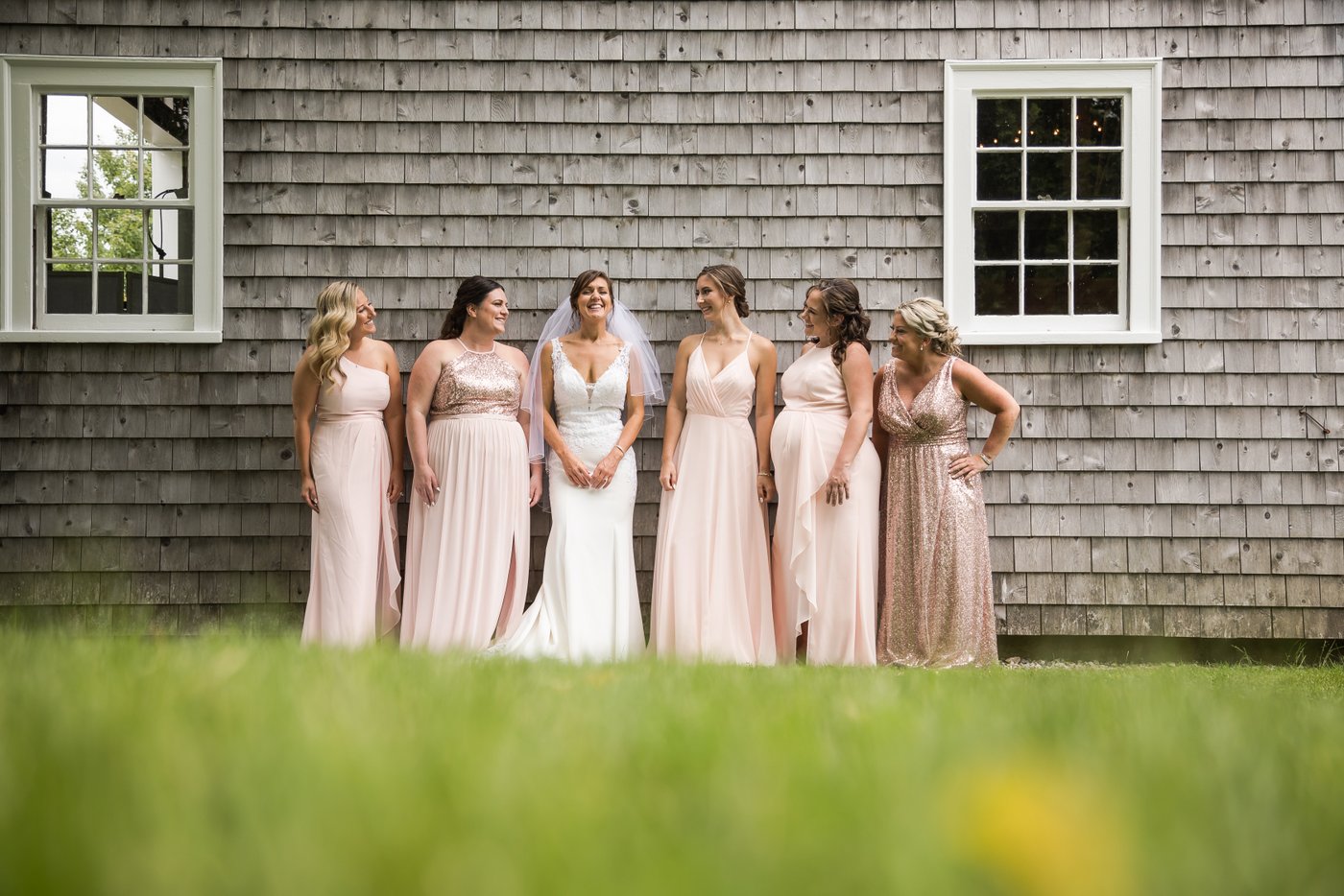 The bride smiles as her bridesmaids watch her as they stand against a rustic gray cabin during a Massachusetts barn wedding