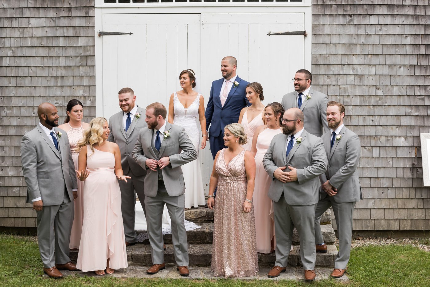 The bride, the groom, and their wedding party stand against a rustic gray barn with white doors during a Massachusetts barn wedding, photographed candidly by Massachusetts wedding photographers Spagnolo Photography