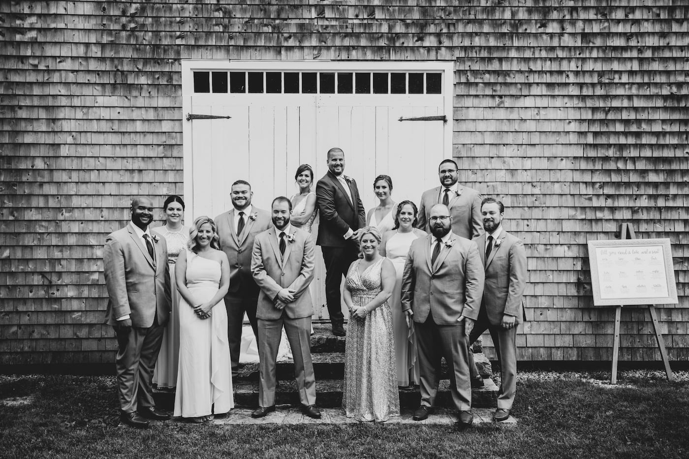 A black and white photo of the bride, the groom, and their wedding party standing against a rustic gray barn with white doors during a Massachusetts barn wedding, photographed by Massachusetts wedding photographers Spagnolo Photography