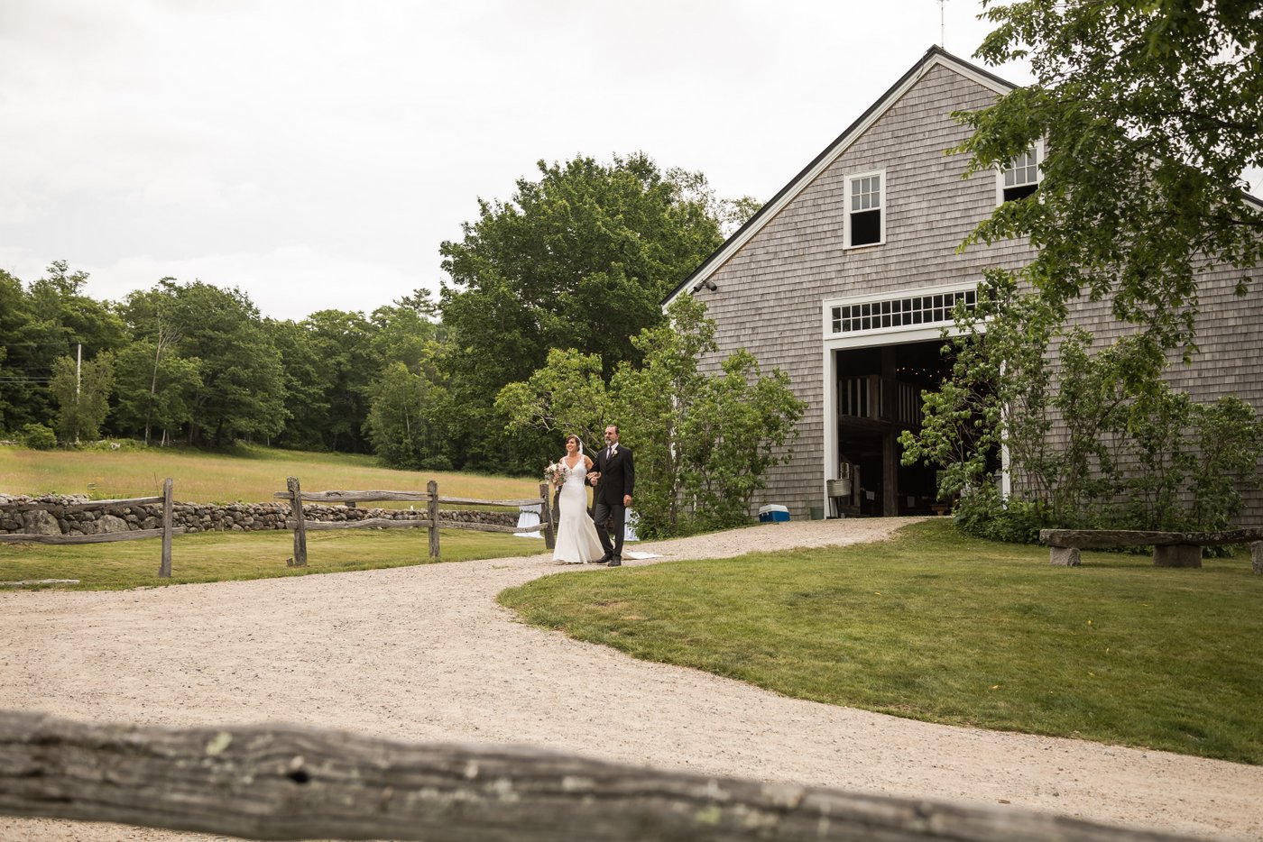 The bride and her father emerge from the barn and begin their processional toward the altar at a barn wedding in Massachusetts