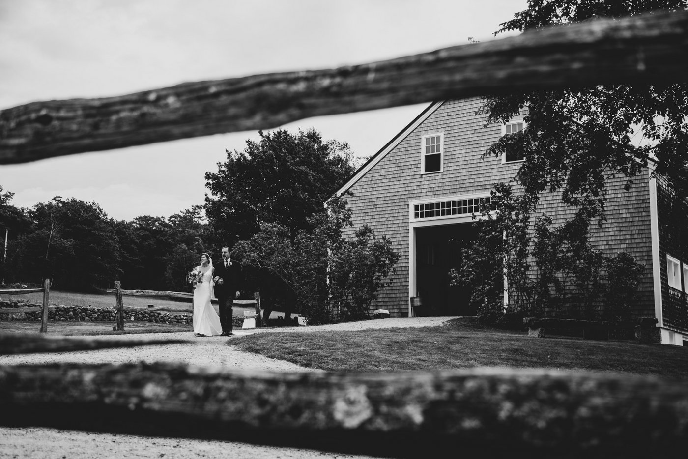A black and white photo of the bride and her father walking out of the barn and toward the altar at a barn wedding in Massachusetts