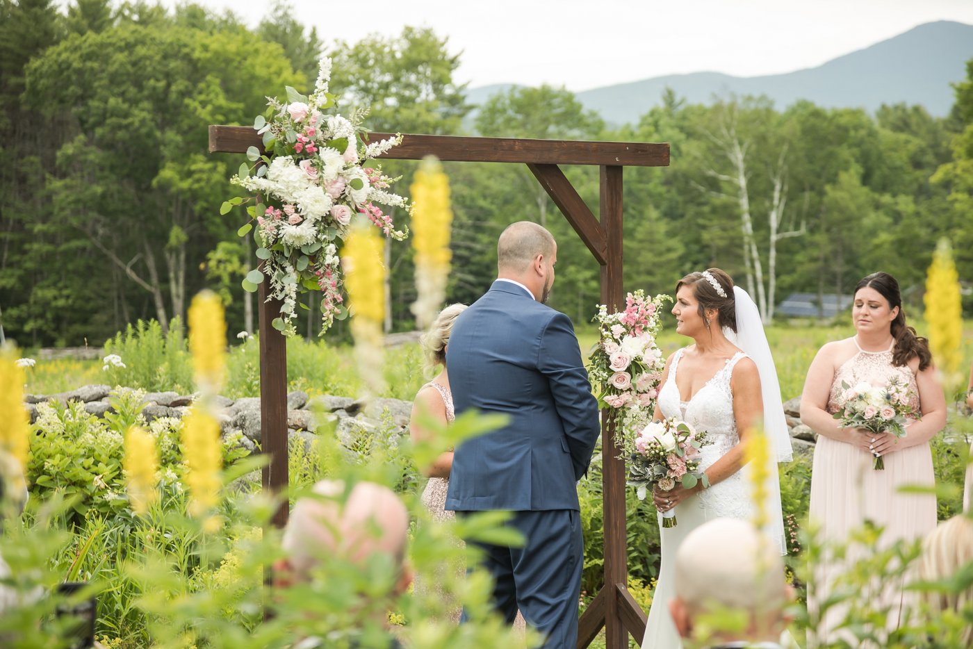 The bride and groom at the wooden altar decorated with flowers, photographed through wildflowers at an outdoor farm wedding in Massachusetts
