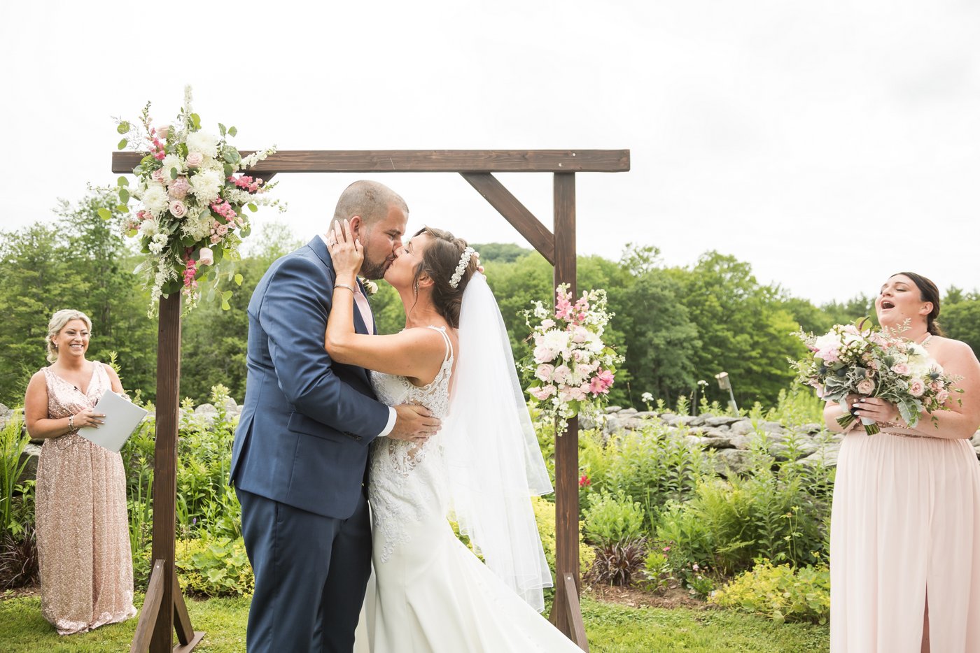 The bride and groom kiss at the wooden altar decorated with flowers during their outdoor rustic farm wedding in Massachusetts