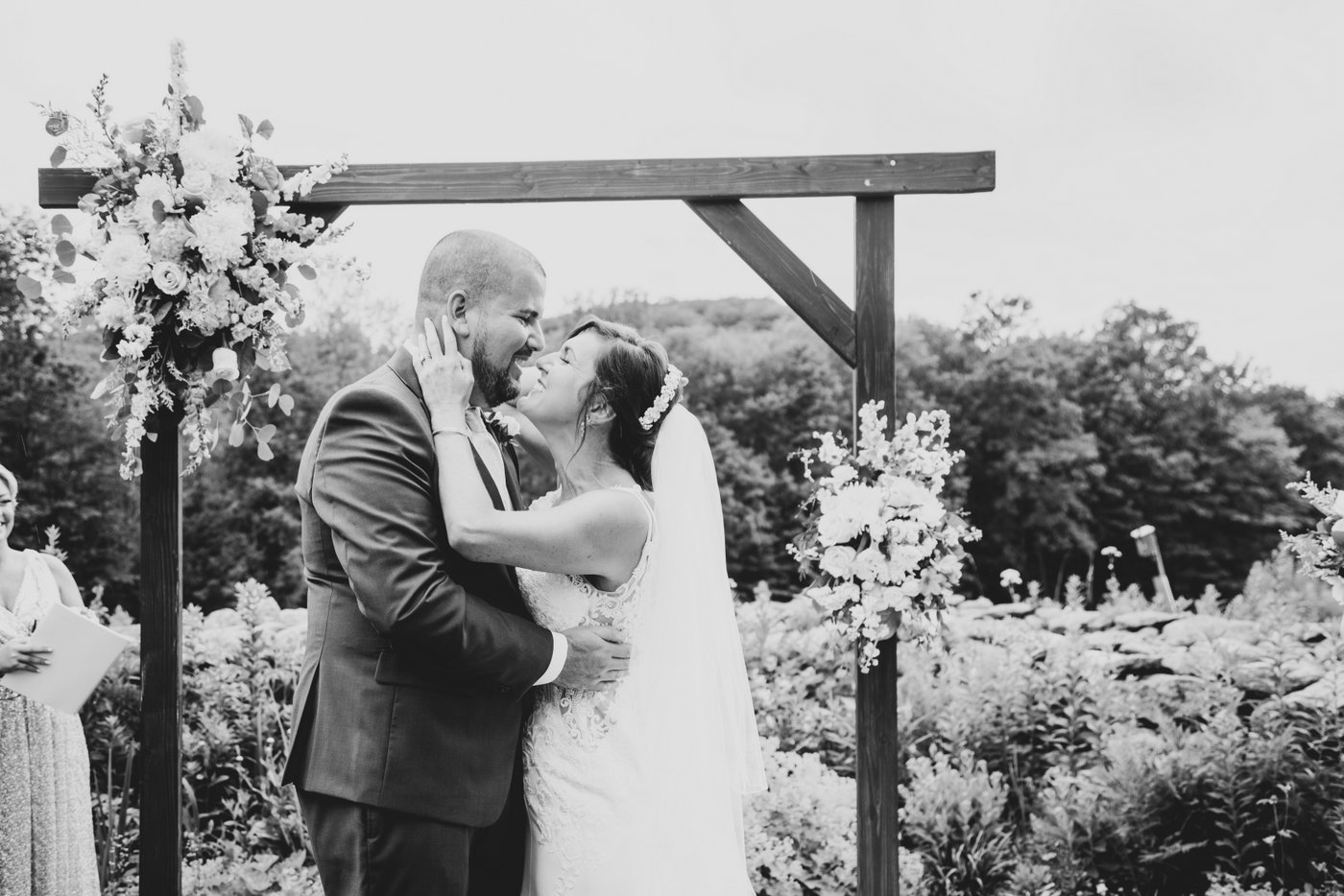 A black and white photo of the bride and groom kissing at the wooden altar decorated with flowers during their outdoor rustic farm wedding in Massachusetts
