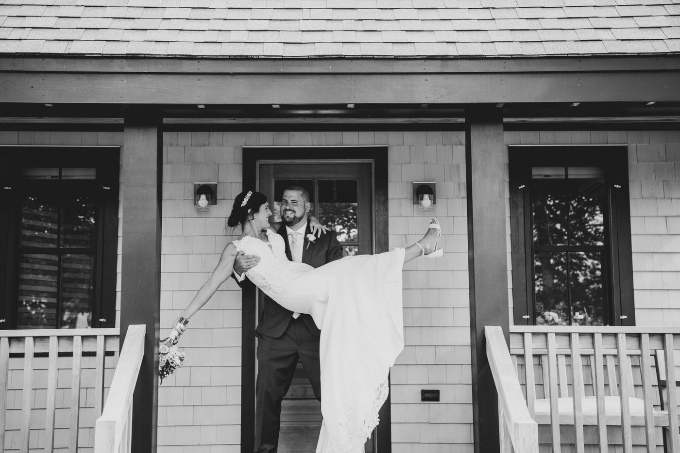 A black and white photo of the groom carrying the bride in front of a small cabin at a barn wedding in Massachusetts