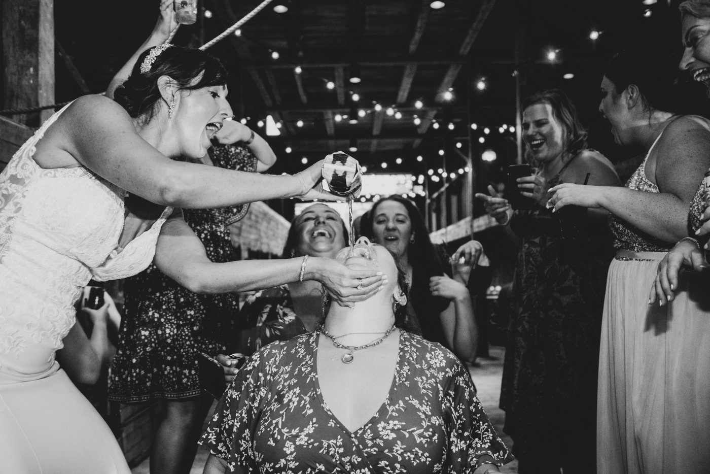 A black and white image of a bride pouring beer in the mouth of a guest as others line up behind her at a barn wedding in Massachusetts