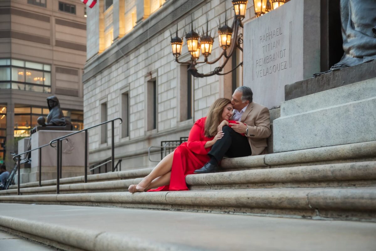 An elegantly dressed couple sits on the steps of the Boston Public Library, photographed on their engagement shoot by Boston engagement photographers Spagnolo Photography in 2025.