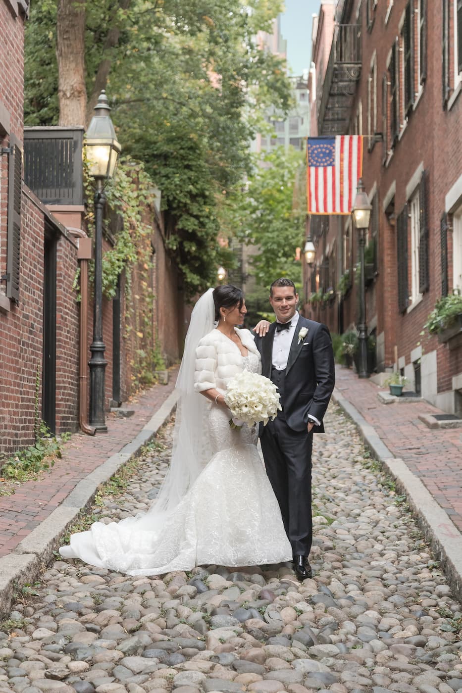 A bride in an elegant white gown and a long veil and a groom in a tuxedo posing for a portrait in the middle of the iconic Acorn Street in Boston, photographed by Boston wedding photographers Spagnolo Photography.