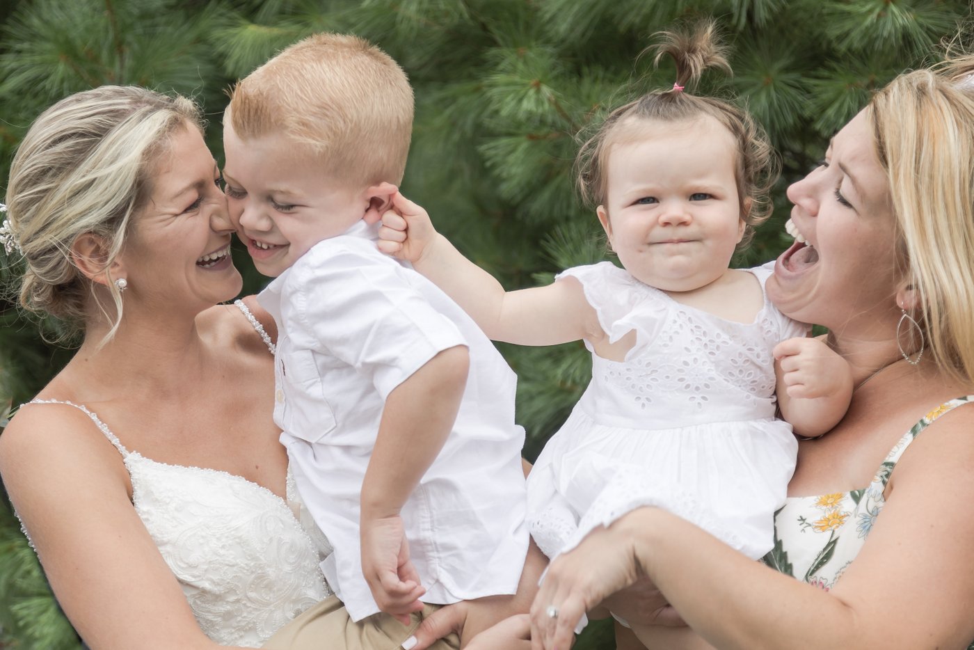 A humorous candid shot of a toddler in her mother's arms pinching the ear of another toddler who's in the arms of a bride