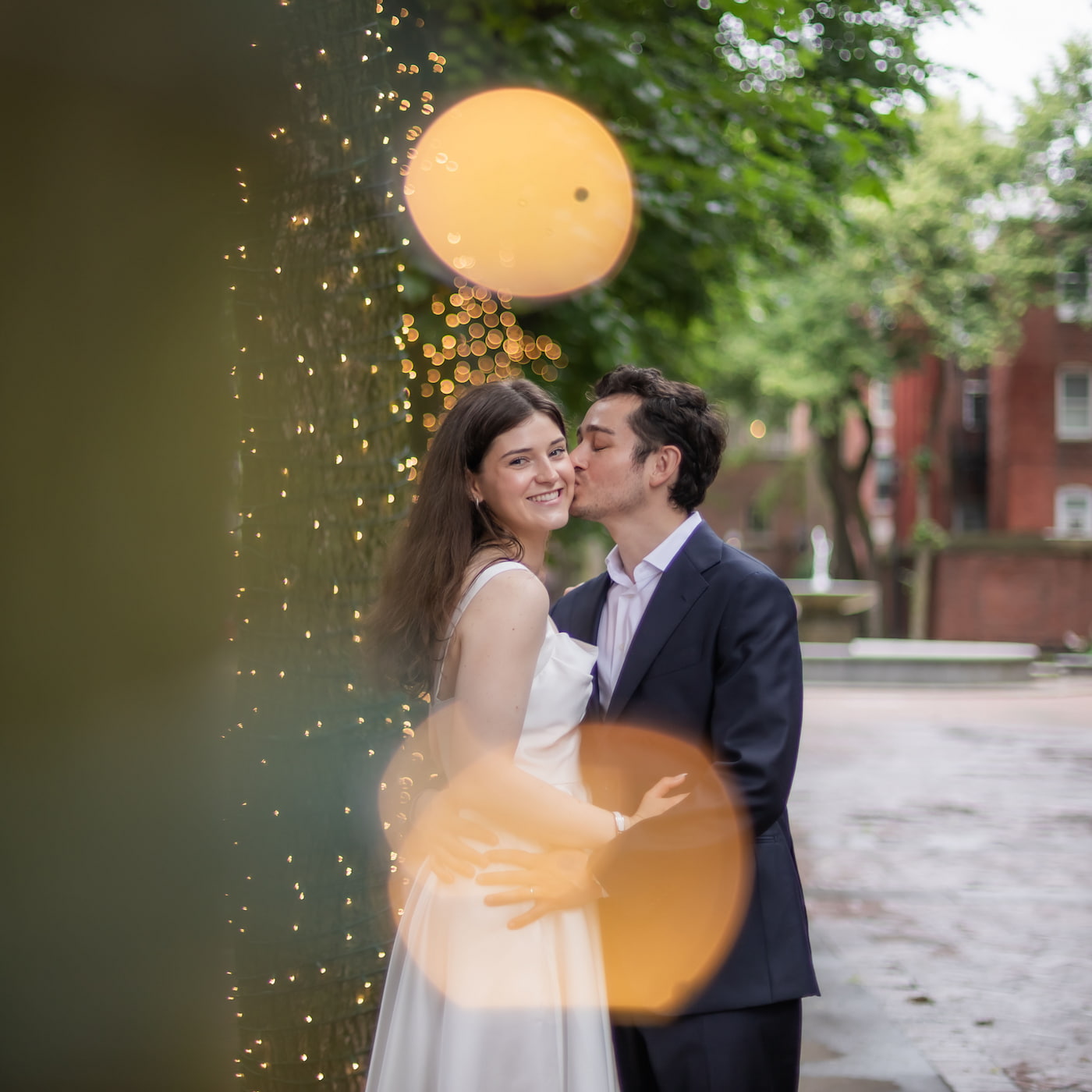 A bride looks at the camera as the groom kisses her cheek, behind them a tree trunk wrapped in sparkling yellow lights that produce a bokeh effect, photographed during a Boston elopement photoshoot by Boston wedding photographers Spagnolo Photography.