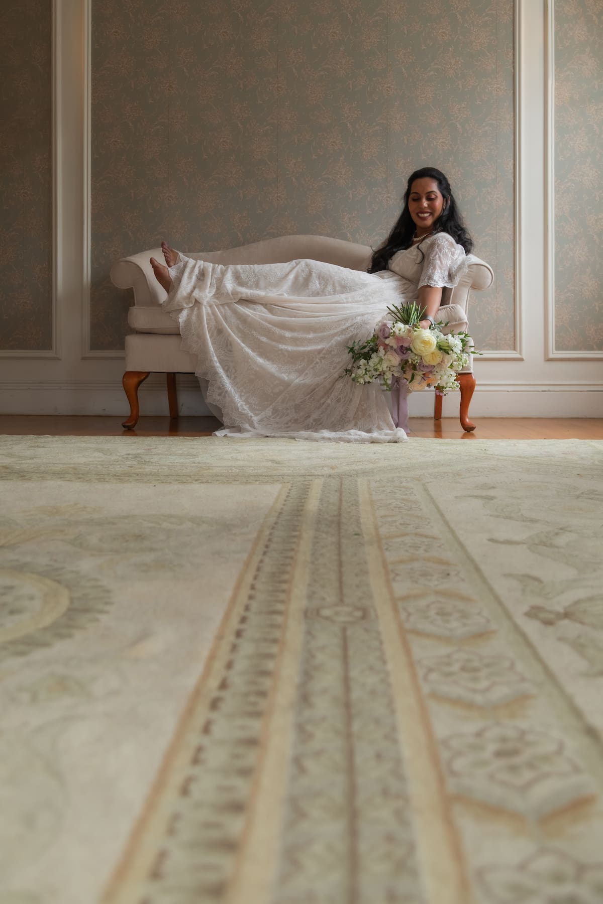An elegant bride in a lace Anthropologie wedding gown reclines on a vintage beige claw-footed couch, holding a large bouquet of pink and white flowers, photographed in an artistic style that incorporates the pattern of the antique rug that leads to her, by Boston wedding photographers Spagnolo Photography during an Endicott Estate wedding in Dedham, MA.