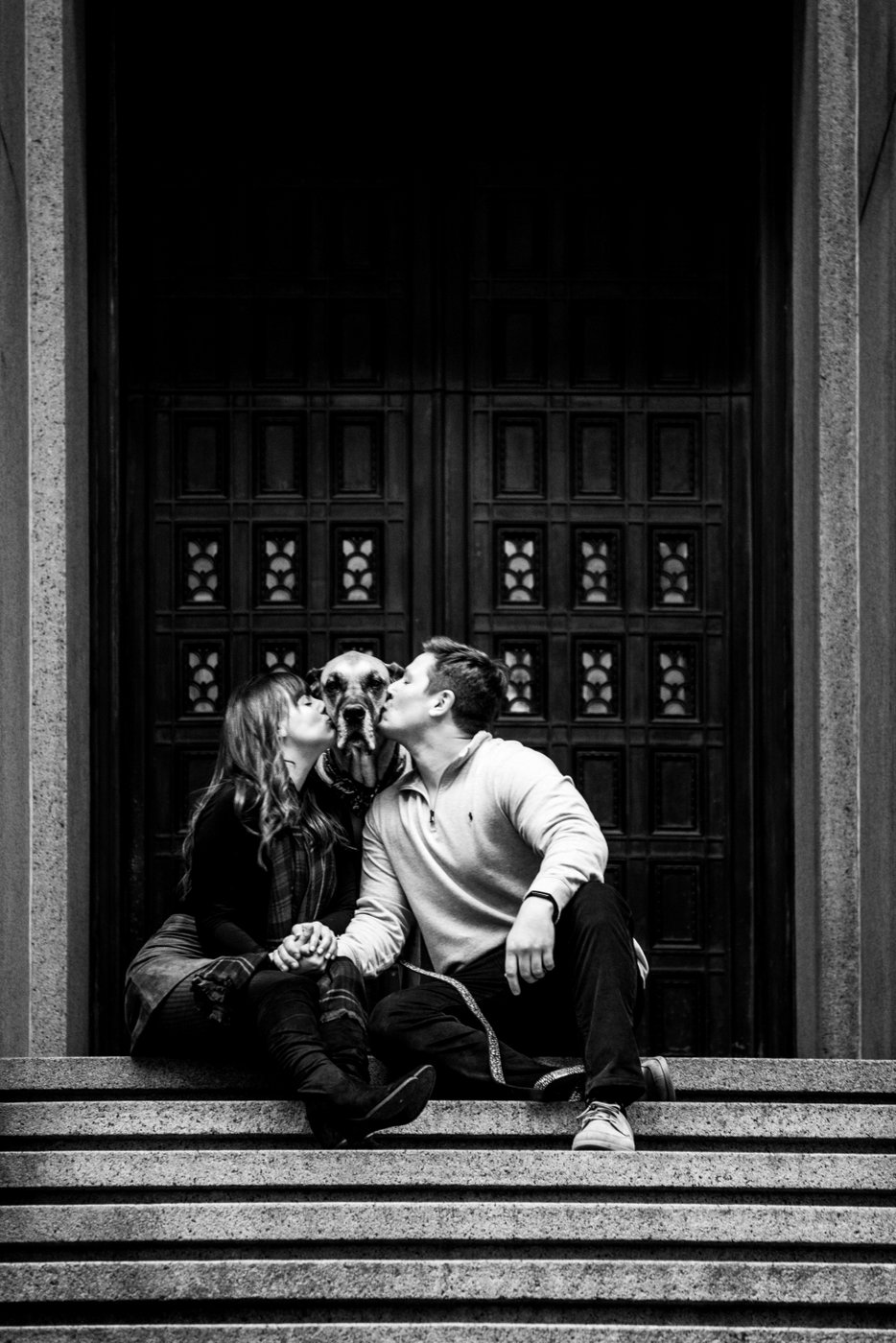 A Boston couples photography portrait of a man and a woman kissing their dog sitting between them on the stairs of a building in Boston, photographed by Boston wedding photographers Spagnolo Photography.