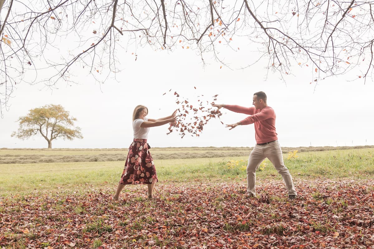 A fun photo from a fall engagement photography session showing a man in a pink sweater and khaki pants and a woman in a white top and a floral skirt throwing leaves at each other in a park, photographed by Boston wedding photographers Spagnolo Photography