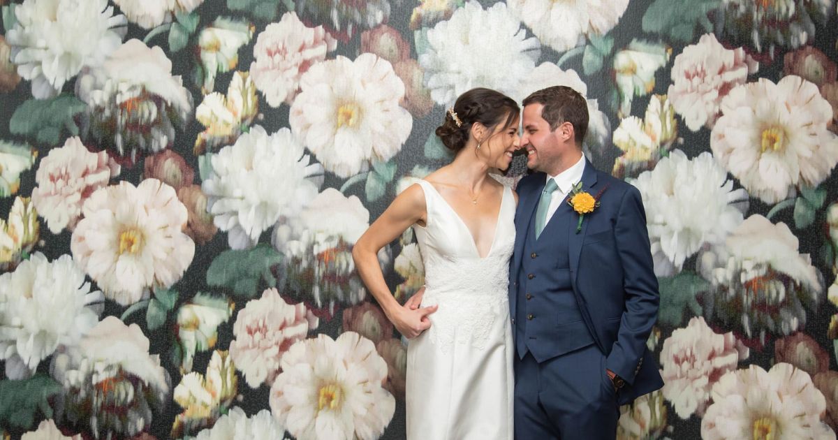 Boston wedding photographers Aaron and Nanore Spagnolo capturing a candid wedding moment between a bride and a groom smiling at each other in front of a wallpaper with large pink and white flowers