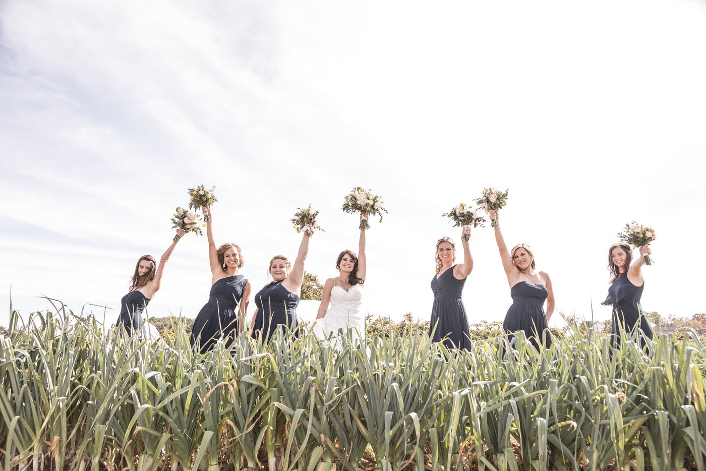 A bride and her bridesmaids standing in the middle of a field at the Barn at Gibbet Hill in Groton raise their flower bouquets, photographed by Boston Wedding Photographers Spagnolo Photography.