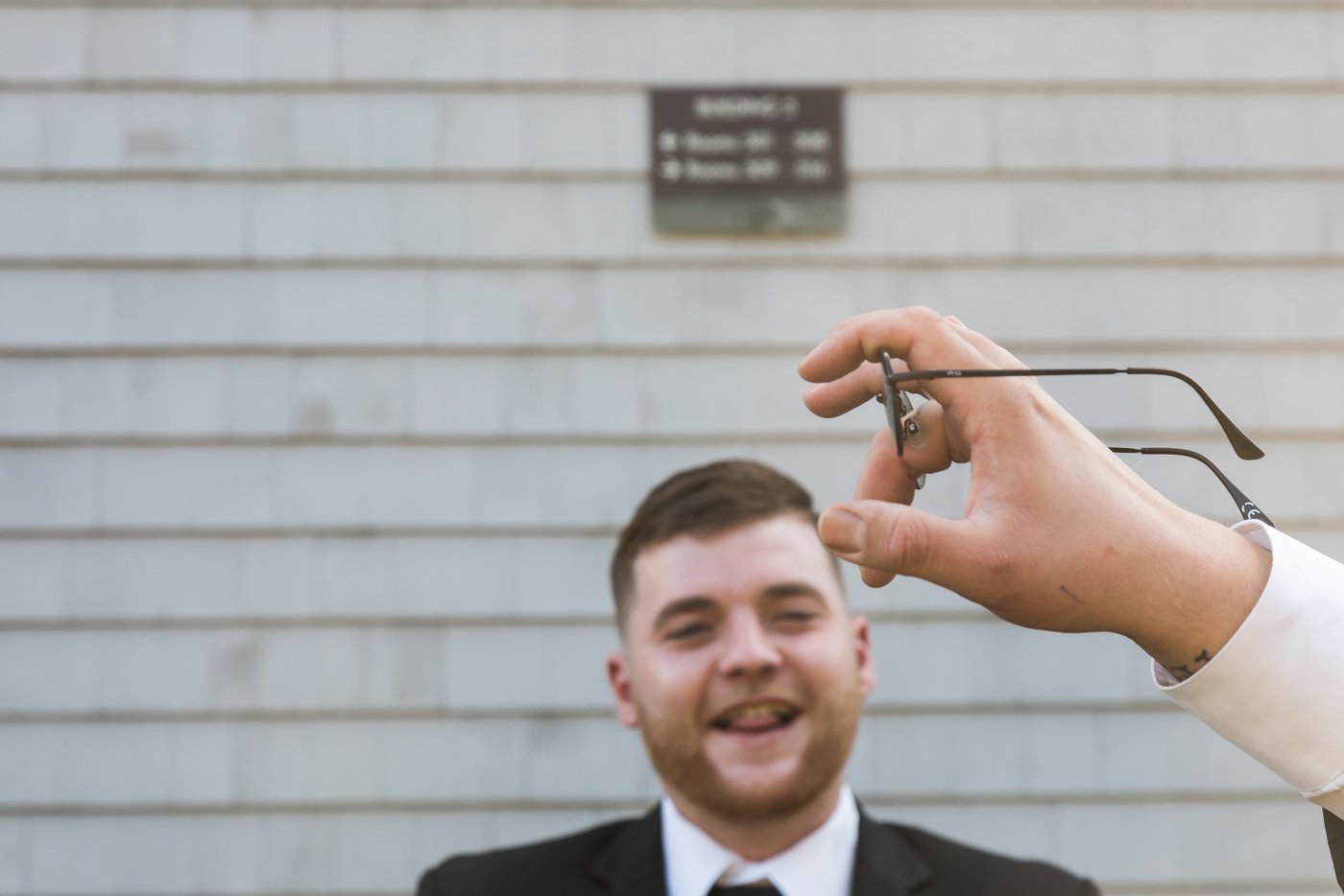 A groomsman squints as another groomsman's hand holds up a pair of glasses, photographed in a candid and funny style by Spagnolo Photography