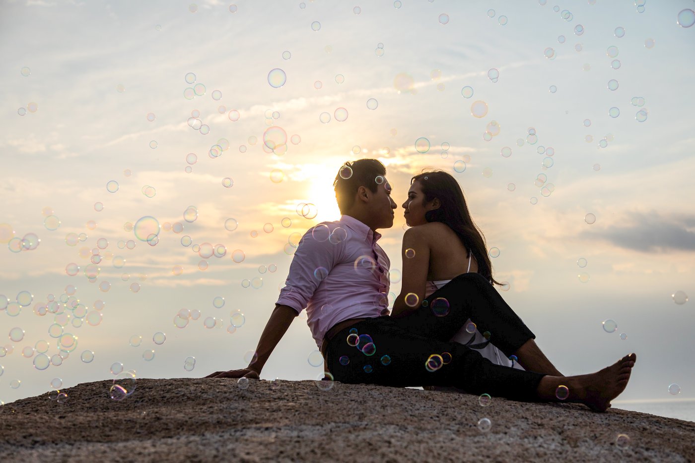 A man and a woman sitting on a rock gaze at each other as the sun sets behind them, photographed at Stage Fort Park in Gloucester during their couples photography session with Boston wedding photographers Spagnolo Photography.