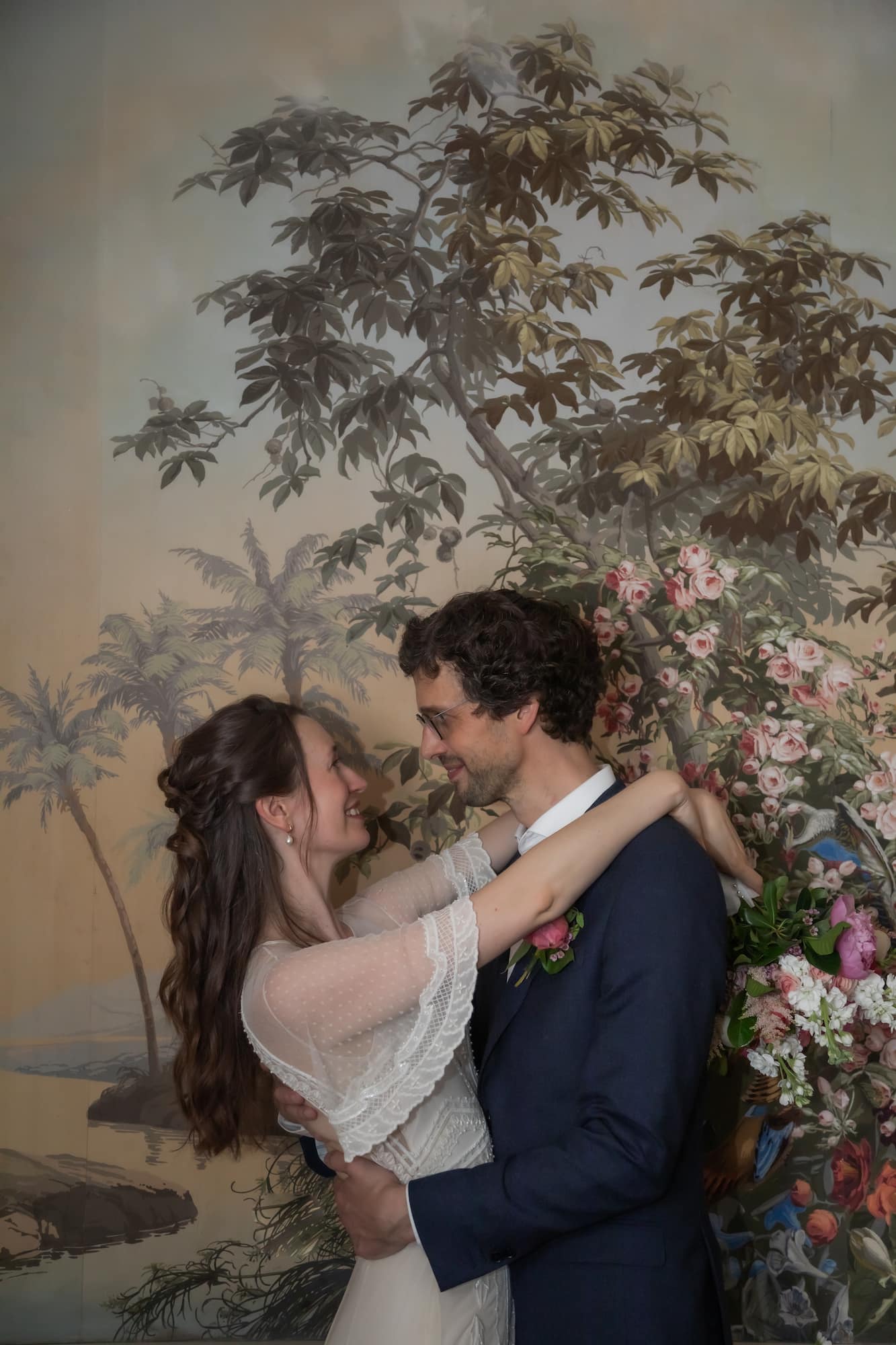 A bride in a lace white dress and a groom in a navy suit hug in front of an antique wallpaper during their Beacon Hill wedding at the King's Chapel Parish House, photographed by Boston wedding photographers Spagnolo Photography in their unique creative style.