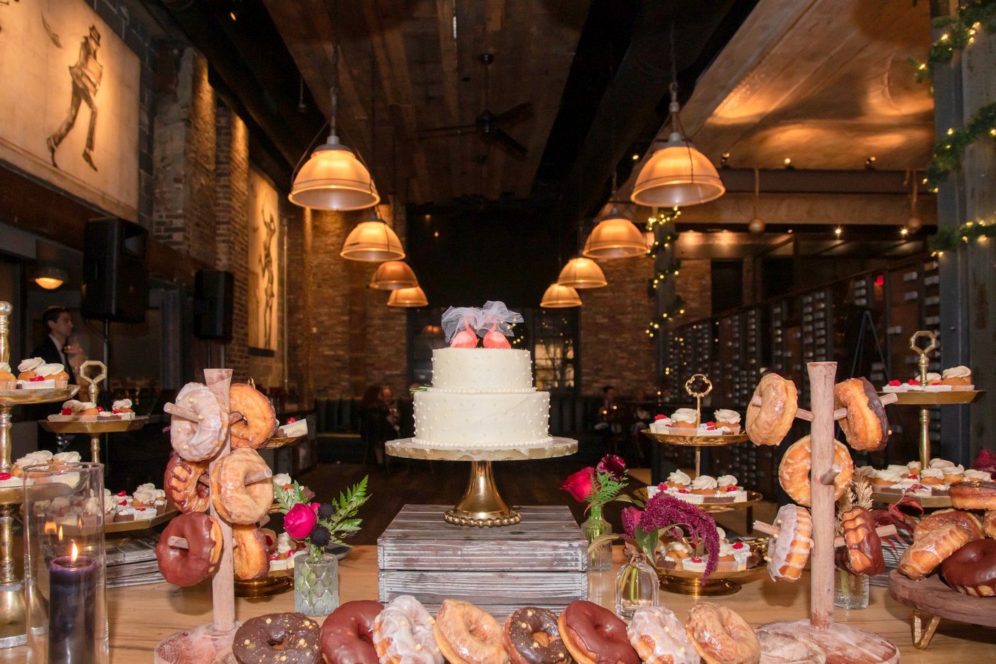 A wedding cake with flamingo toppers, donuts, and cupcakes are piled on a wooden table at The Ledger Restaurant wedding in Salem, MA, photographed in an angle that incorporates the interior design, particularly the light fixtures in a way that maximizes the cinematic effect of the scene, in an artistic style unique to the Boston-based creative photographer duo, Spagnolo Photography.