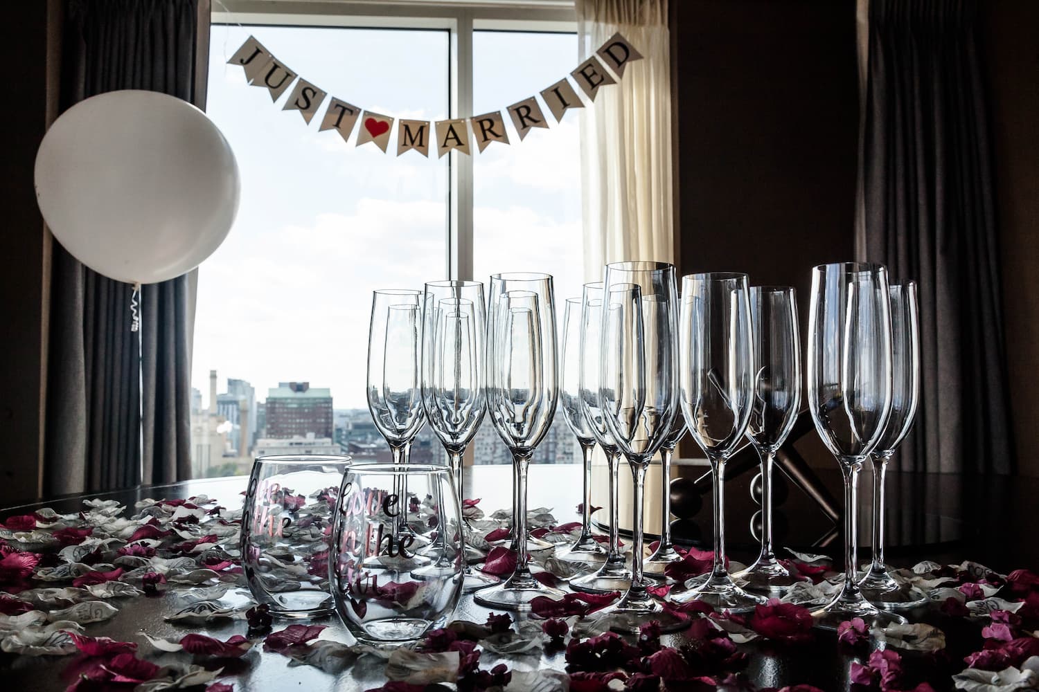 A creative shot of clean empty flute glasses and rose petals on a table, a white balloon hovering by the window, and a banner that reads "Just Married" in the background, photographed at a Liberty Hotel wedding by Boston wedding photographers Spagnolo Photography in their creative style.