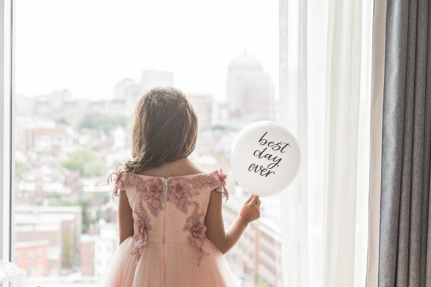 A flower girl stands looks out the window of a bridal suite at the Liberty Hotel in Boston while holding a white balloon that reads "Best Day Ever" while the bride gets ready in another room