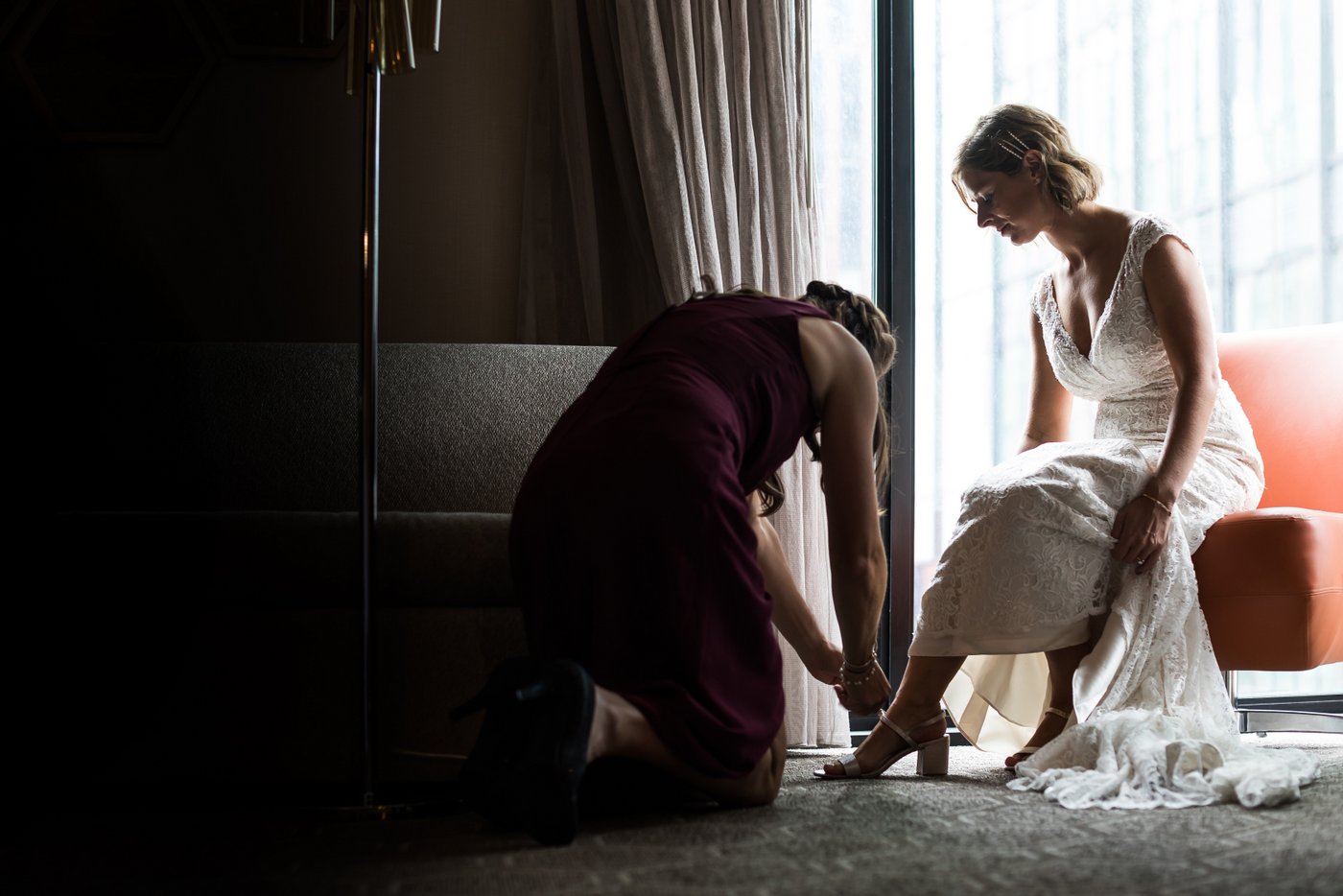 A bridesmaid helps buckle the bride's shoe in a moodily lit hotel room at the Marriott in Cambridge