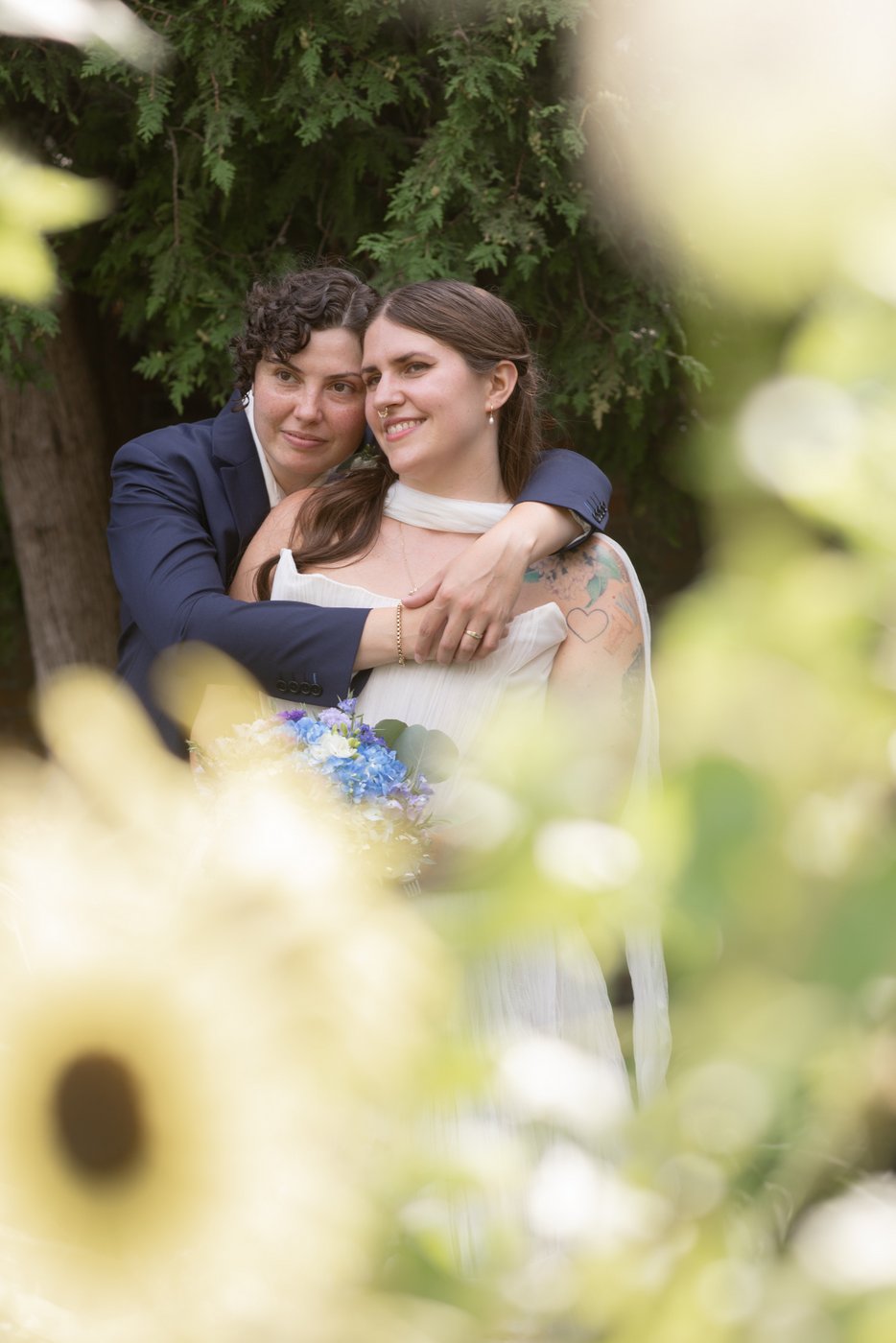 A wedding portrait of an LGBTQ couple hugging in a garden, shot through a bed of flowers by creative wedding photographers Spagnolo Photography during a wedding portrait session at the garden of Ropes Mansion in Salem, MA.