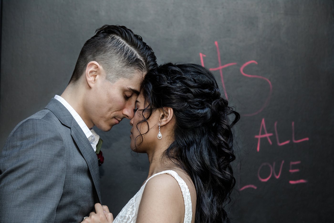 Artistic Boston wedding photographers Spagnolo Photography capturing a candid photo of a bride and a groom leaning their foreheads together, their eyes closed, as they take a moment to themselves during their wedding at Sarma Restaurant in Somerville, MA, the words "It's All Love" are written on the black wall behind them.