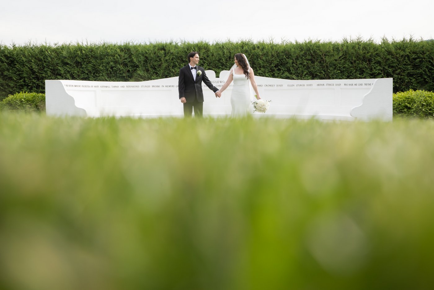 A bride and a groom hold hands and look at each other as they stand in front of the Whispering Bench during their Tanglewood wedding, photographed from a low angle and through freshly cut grass by artistic wedding photographers Spagnolo Photography, known for close attention to photo composition and geometry.