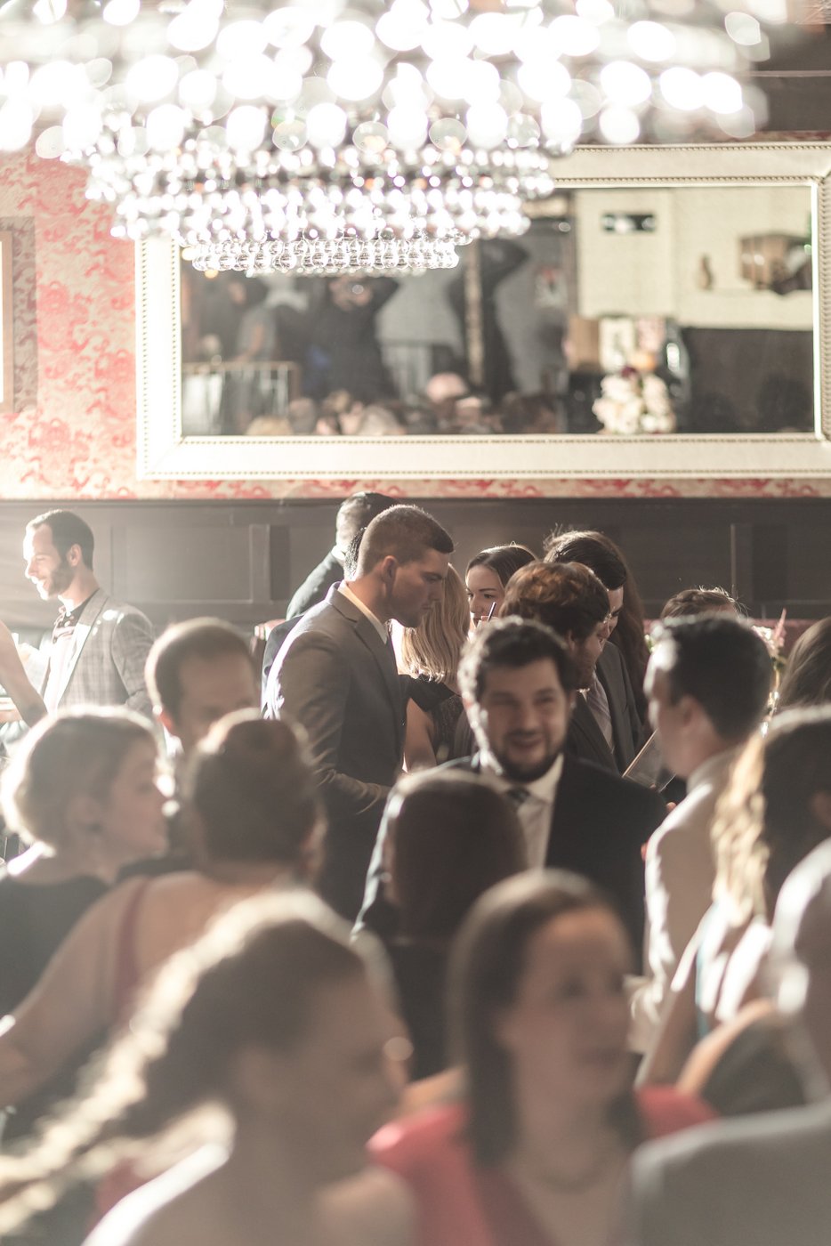 An artistic candid photo of wedding guests mingling under a chandelier during cocktail hour at a wedding reception at the Wink and Nod Speakeasy in Boston.