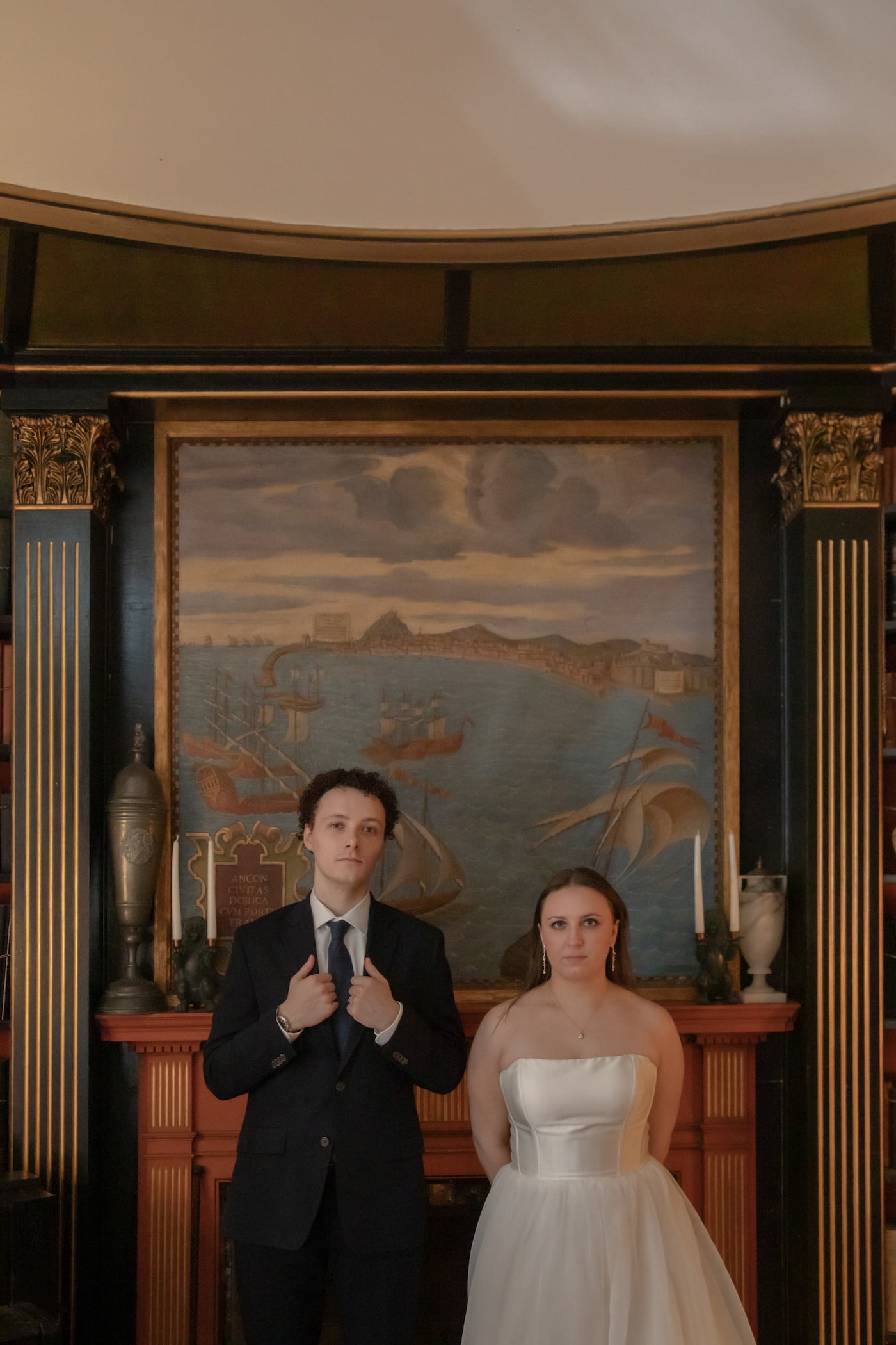 A portrait of a bride and a groom looking at the camera with deadpan expressions in the library of Hammond Castle Museum on their wedding day, photographed by candid Boston wedding photographers Spagnolo Photography.
